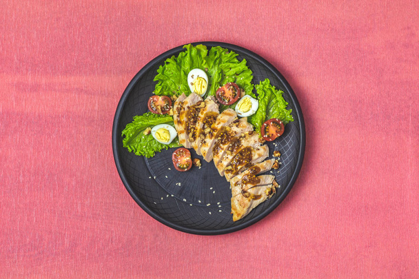 Delicious salad, chicken with honey and mustard souse, lettuce, quail eggs, cherry tomatoes and sesame in black ceramic plate, top view, copy space, hawaiian, asian food, living coral table surface - Photo, Image