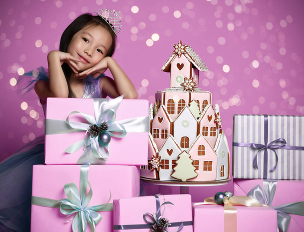 Little asian girl in crown and purple dress with many present gift boxes and ginger cookie cake birthday celebration  - Photo, Image