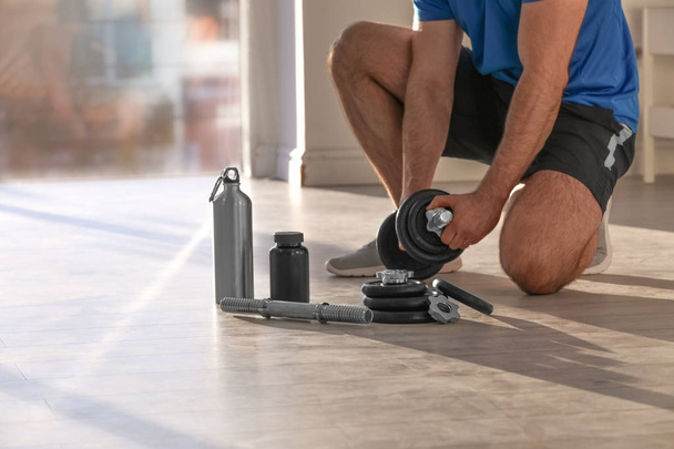 Male athlete assembling adjustable dumbbell near bottle, parts and jar of protein on floor indoors - Foto, afbeelding