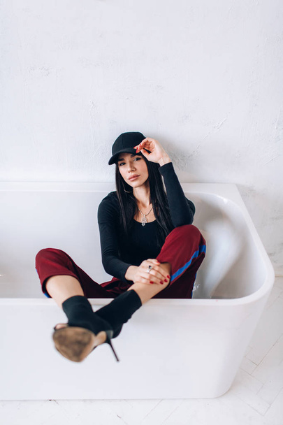 Portrait of a brunette girl in a cap and sportswear. Natural light. Red sweatpants, black top, black cap. - Photo, image