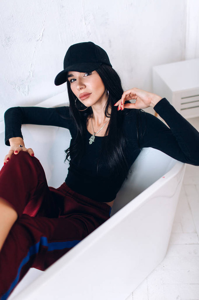 Portrait of a brunette girl in a cap and sportswear. Natural light. Red sweatpants, black top, black cap. - Photo, Image