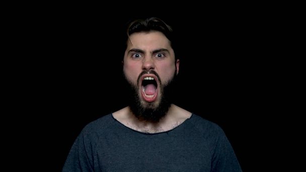 Портрет рыкающего красавца с бородой, стоящего и кричащего с большим открытым ртом, изолированного на черном фоне. Молодой сердитый бородатый мужчина кричит и кричит
 - Фото, изображение