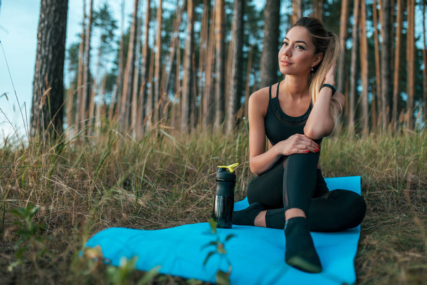 Girl resting in the forest on a yoga mat. Near a bottle of water. camping. Woman doing sports in the summer in the park in the fresh air. - Фото, изображение