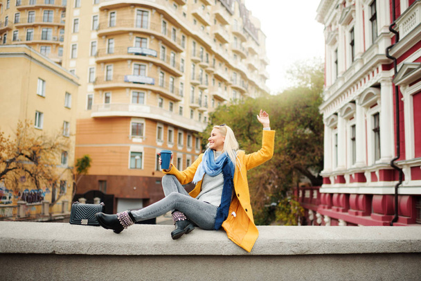 Stylový šťastná Mladá blondýnka v zářivě žlutém plášti drží kávu s sebou. Chůzi na ulici. Portrét mladé ženy veselý při přerušení práce. Životní styl koncept  - Fotografie, Obrázek