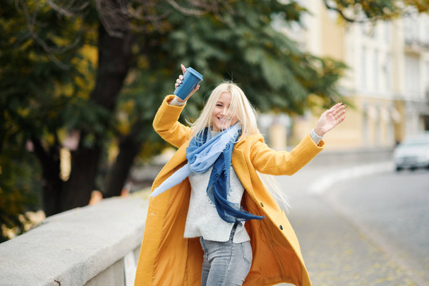 stylische junge blonde Frau in leuchtend gelbem Mantel hält Coffee to go. zu Fuß auf der Straße. Porträt einer jungen, fröhlichen Frau in der Arbeitspause. Lifestylekonzept  - Foto, Bild