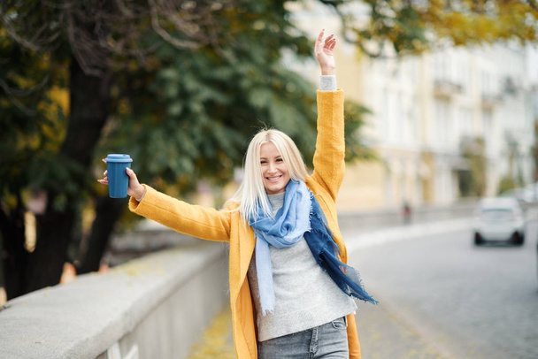 stylische junge blonde Frau in leuchtend gelbem Mantel hält Coffee to go. zu Fuß auf der Straße. Porträt einer jungen, fröhlichen Frau in der Arbeitspause. Lifestylekonzept  - Foto, Bild