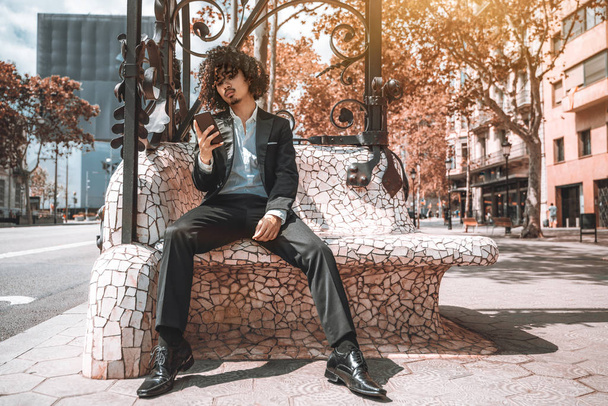 An arrogant bearded Asian guy with curly hair and in a business suit is using his smartphone to see incoming messages while sitting outdoors on the street bench between the road and the sidewalk - Фото, изображение