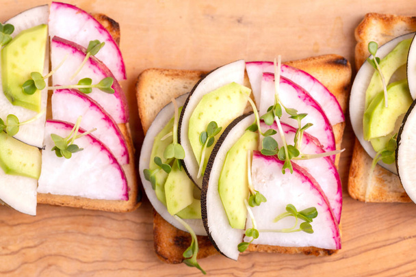 Tasty wheat toasts with radish, avocado and sprouts on wooden cutting board. Dark concrete background. Simple healthy breakfast. Copy space. Top view - Φωτογραφία, εικόνα