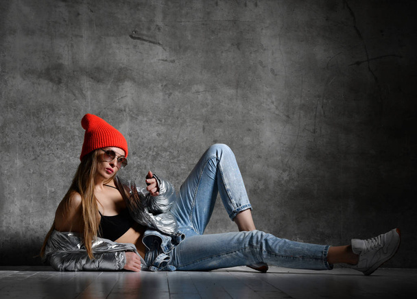 Young sport woman after workout exercise sitting point finger in silver sexy thin down puffer jacket blue jeans red hat  - Foto, Imagen