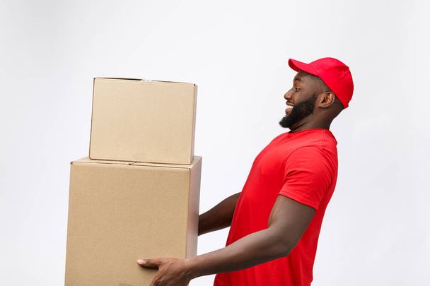 Концепция доставки - Вид сбоку Портрет счастливого афроамериканского курьера в красной ткани с коробкой в руках. Изолирован на сером фоне. Копировальное пространство - Фото, изображение