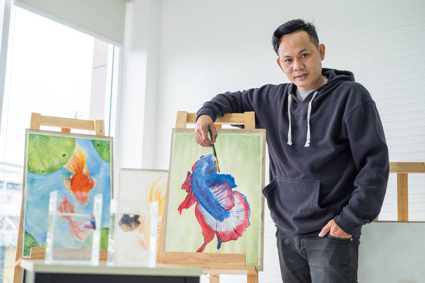Nuori mies aasialainen maalarit seistä esitellä hänen taiteellista työtä. Kuva kauniista kalasta inspiraation avulla. Antaa arvokkaan panoksen
. - Valokuva, kuva