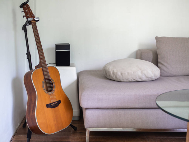 Akustik gitar ve hoparlör beyaz yan masada kanepe yastık ve beyaz rahat oturma odasında ahşap zemin üzerinde yuvarlak cam masa ile yakın. Söz yazarı kavramı. - Fotoğraf, Görsel