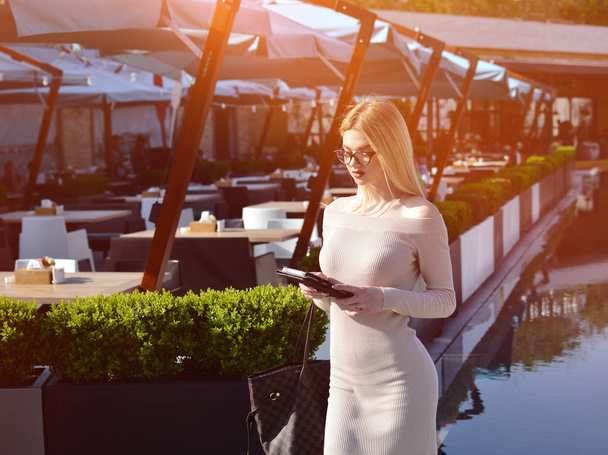 Attractive blonde girl is pulling a smartphone in the background of a restaurant with a swimming pool. People in the work process. Online work. - Foto, immagini