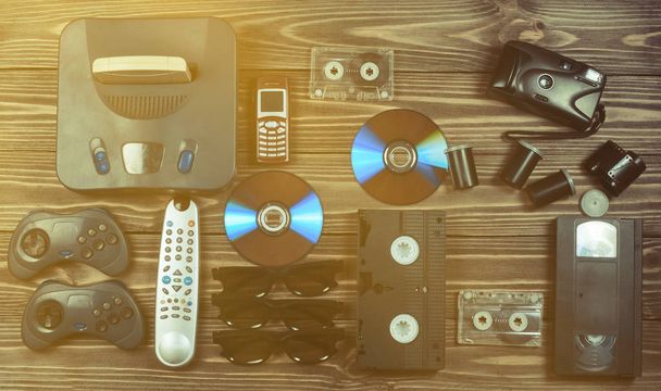 Vintage media and information technology. Entertainment 90s. Game console, gamepads, disks, audio cassettes, video cassettes, phone, film camera on a wooden table. Top view. Flat lay. - Photo, Image