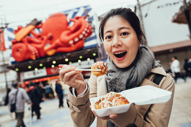 young asian woman traveler trying takoyaki face camera showing by chopsticks. tourist excited open mouth eating tasty octopus balls. delicious japanese street food. - Foto, afbeelding