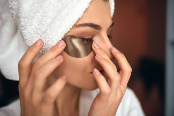 Closeup indoor portrait of beautiful woman applying eye patches on the area under the eyes from wrinkles and dark circles. Attractive female in white bathrobe making beauty treatments at home.  - Foto, Imagen