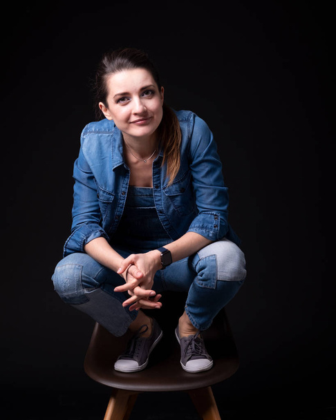 brunette girl in blue denim jacket and pants stands leg up on chair close up on black background - Foto, afbeelding
