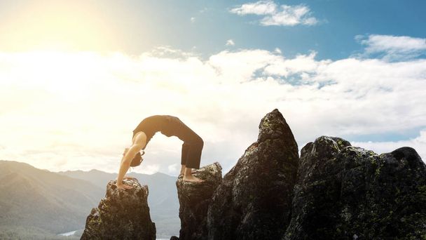 man doing complex Yoga exercise headstand. Amazing Yoga landscape in beautiful mountains. Dangerous stunts traceur standing on his hands on the edge of a cliff. - Photo, Image