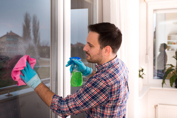 Красивый молодой человек моет окна тряпкой и баллончиком дома
 - Фото, изображение