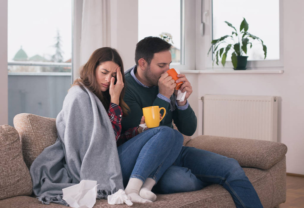 Young couple sitting on sofa covered with blanket, drinking tea and blowing nose. Cold and flu season  - Photo, Image