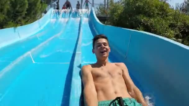 Happy young man laughing and taking a selfie on a water flume in an aqua-park                        Cheerful view of sportive brunet man in long shorts taking a selfie, laughing happily and riding on a water chute in a Turkish aqua-park in summer - Πλάνα, βίντεο