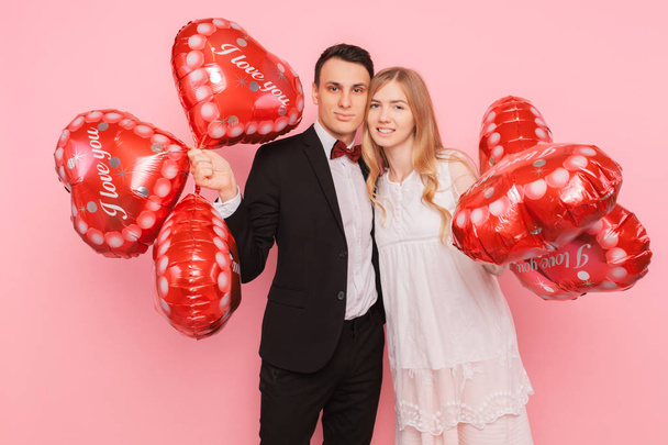 A loving couple, a man and a woman, holding heart-shaped balloons, in a studio on a pink background - Fotó, kép