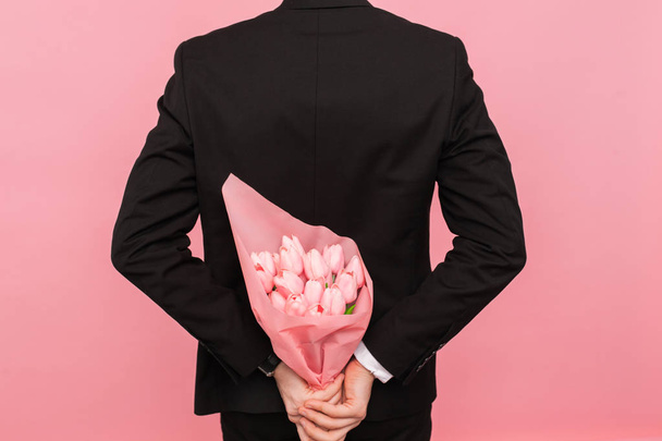 A man holds a bouquet of tulips behind his back, a surprise for a woman, on a pink background, the concept of love and relationships - 写真・画像
