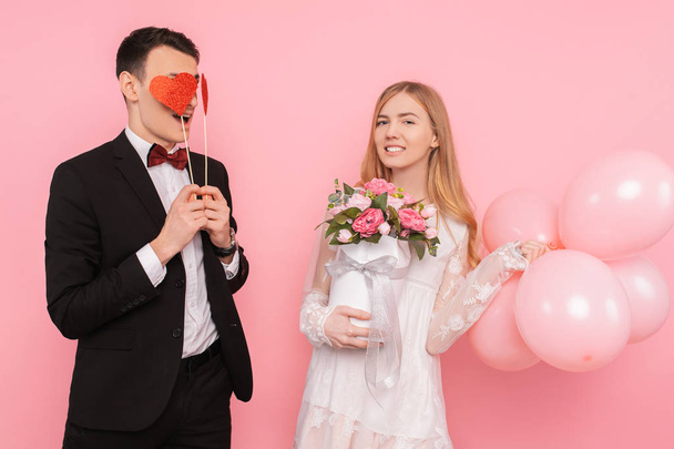 A loving couple, a man holds two paper hearts in his eyes, and a woman holding a bouquet of flowers and balloons in the studio on a pink background - Fotografie, Obrázek
