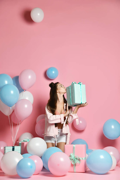 Portrait of young woman in birthday party holding gift box with gift present on pastel pink background with colorful air baloons.  - Foto, afbeelding
