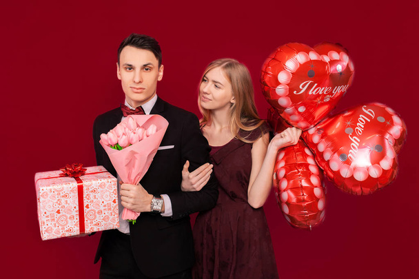 A man and a woman give each other gifts, hold gift boxes and balloons, Valentine's, in the studio on a red background - Фото, изображение