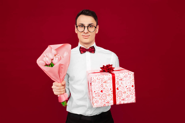 man, wearing glasses, with a gift and a bouquet of flowers, on a red background, the concept of women's day - Фото, зображення