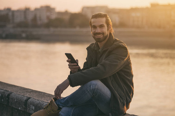 Handsome modern businessman using phone while sitting by the river.Toned image. - 写真・画像