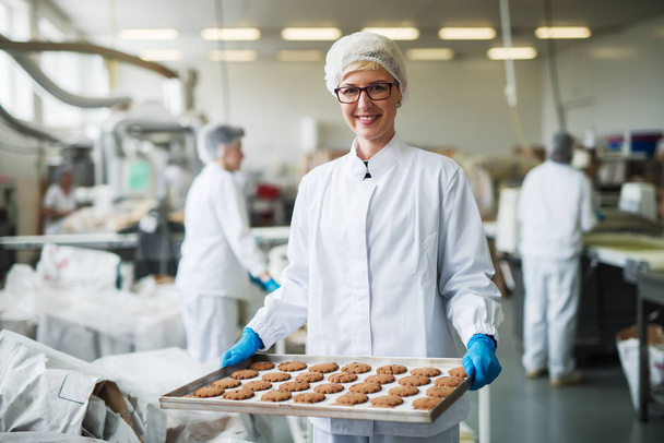 Γυναίκα εργαζόμενη στέκεται και πλατώ με τα cookies. Εσωτερικό εργοστάσιο τροφίμων. - Φωτογραφία, εικόνα