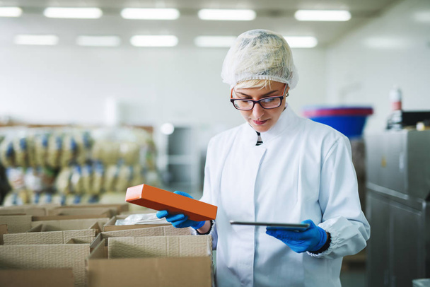 Trabajadora que sostiene la caja de galletas y usa la tableta para controlar el stock mientras está de pie en la fábrica de alimentos
. - Foto, Imagen