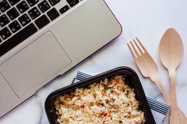 Νόστιμο τηγανητό ρύζι στο κουτί γεύμα στα napery με ξύλινο κουτάλι και πιρούνι και υπολογιστή φορητό υπολογιστή στο χώρο εργασίας για έτοιμα για κατανάλωση και την έννοια των τροφίμων - Φωτογραφία, εικόνα