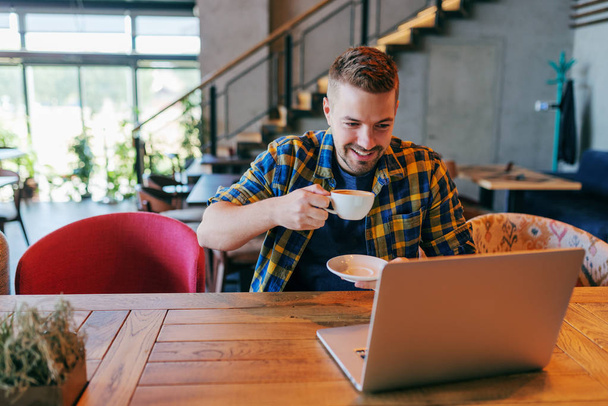 Lachende Kaukasische mannelijke freelancer koffie drinken en laptop kijken zittend in een café. Zelf concept in dienst. - Foto, afbeelding