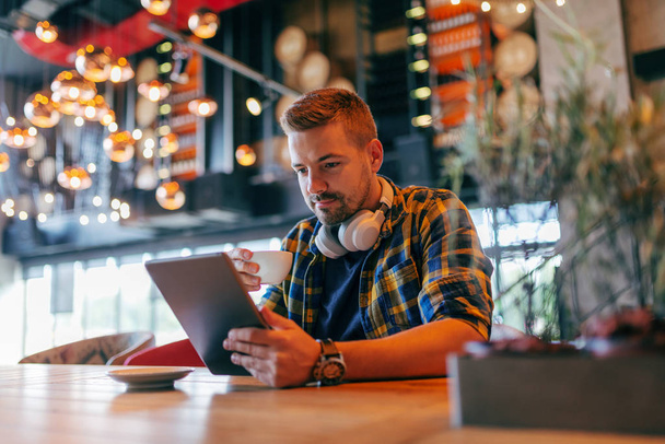 Freelancer jovem caucasiano com fones de ouvido em torno do pescoço usando tablet e beber café enquanto está sentado na cafetaria
. - Foto, Imagem