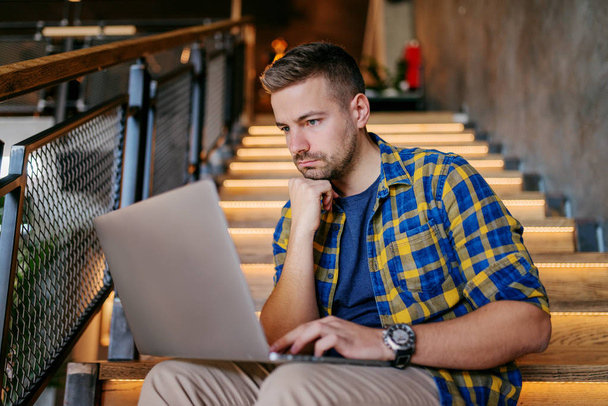 Freelancer barbudo caucasiano usando laptop e segurando a mão no queixo enquanto sentado em escadas na cafetaria. Laptop no colo
. - Foto, Imagem