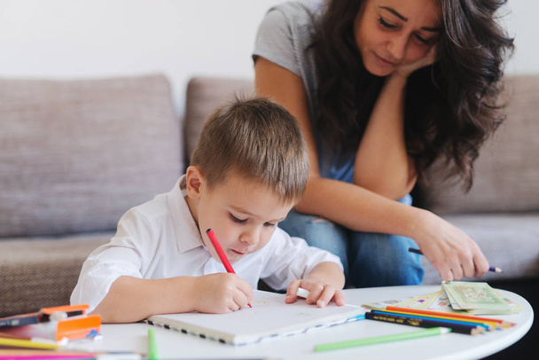 Мальчик рисует карандашами, пока его мать сидит на диване и смотрит на него. Сосредоточься на ребенке. Интерьер дома
. - Фото, изображение