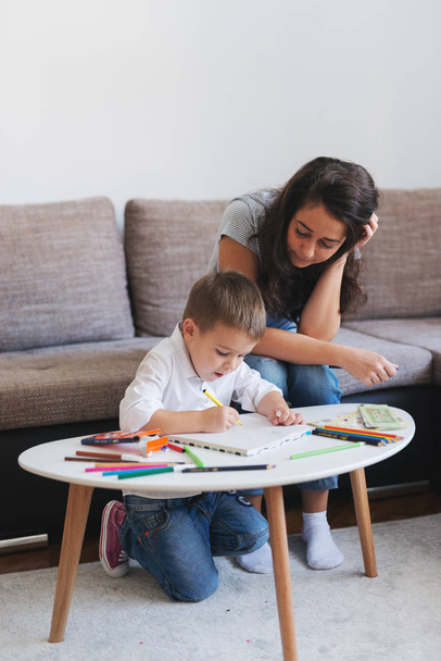 Little boy drawing with crayons while his mother sitting on the couch and looking at him. Home interior. - 写真・画像