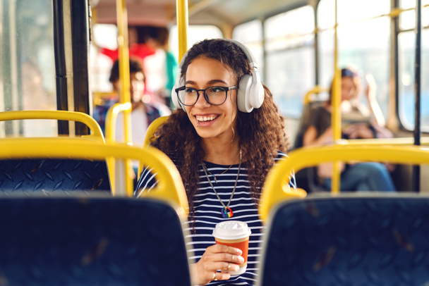 Девушка сидит в автобусе, пьет кофе, слушает музыку и смотрит через окно
. - Фото, изображение