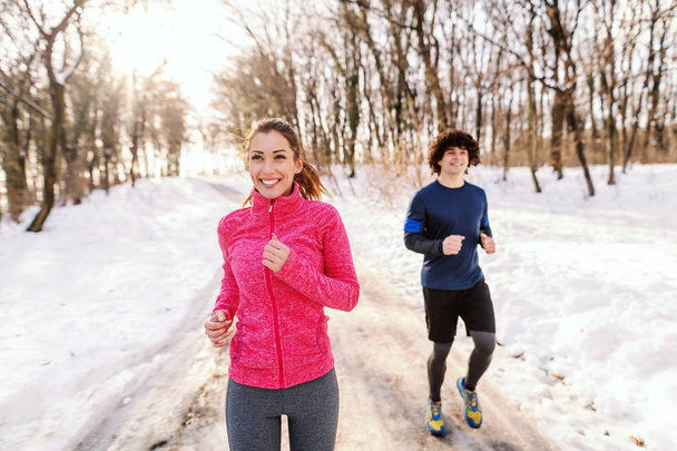 Happy Caucasian friends in sportswear running outdoors. Wintertime, snow all around. Healthy lifestyle concept. - Photo, Image