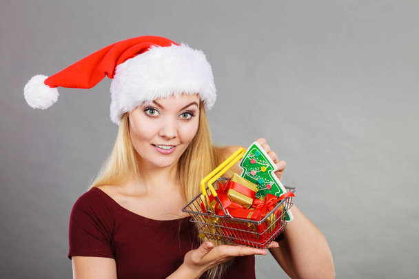Xmas, seasonal sales, winter celebration concept. Happy woman wearing Santa Claus helper hat holding shopping basket cart with little christmas tree and gifts inside. - Foto, afbeelding