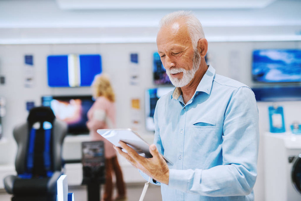 Hombre mayor con seria tableta de pruebas faciales que quiere comprar mientras está de pie en la tienda de tecnología
. - Foto, imagen