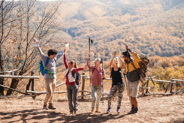 Веселая небольшая группа туристов, стоящих на поляне с поднятыми руками осенью. На заднем плане горы и лес
. - Фото, изображение