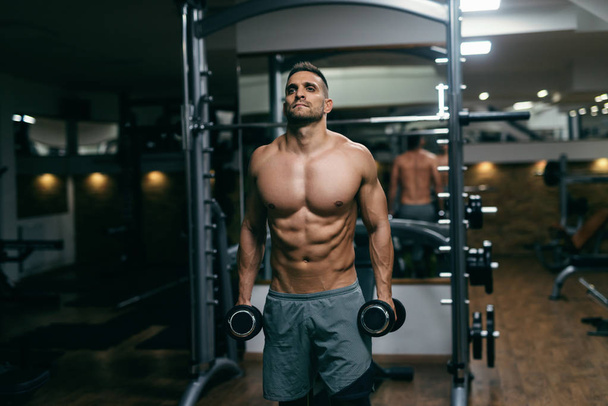 Portrait of shirtless muscular man standing and holding dumbbells in hands. - Фото, зображення