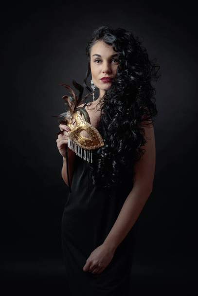 Young beautiful woman with long curly hair in evening gown. Portrait of attractive brunette in evening dress with carnival mask. - Foto, Imagen