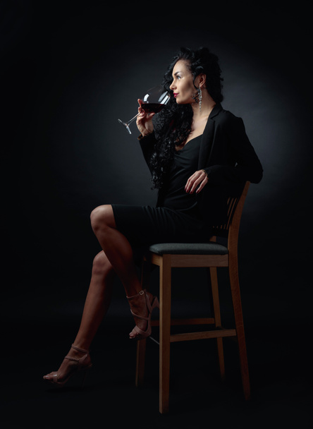 Young beautiful woman with long curly hair in black. Portrait of attractive brunette with glass of red wine on a black bacground. - Fotoğraf, Görsel
