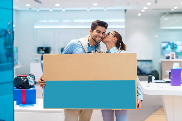 Feliz pareja multicultural sosteniendo la televisión desempaquetada mientras está de pie en la tienda de tecnología. Mujer besándose hombre en la mejilla
. - Foto, imagen