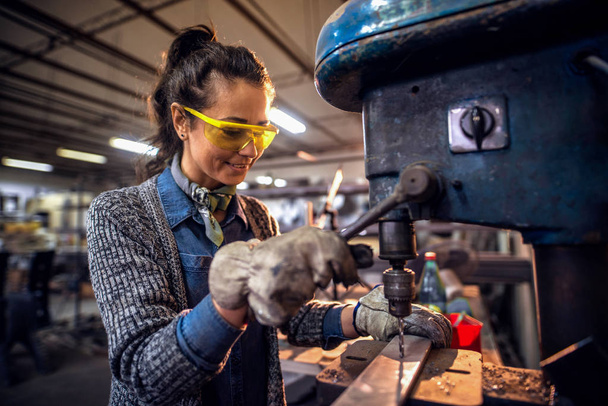 Женщина с помощью вертикальной сверлильной машины, чтобы сделать отверстие в металле, стоя в мастерской
. - Фото, изображение